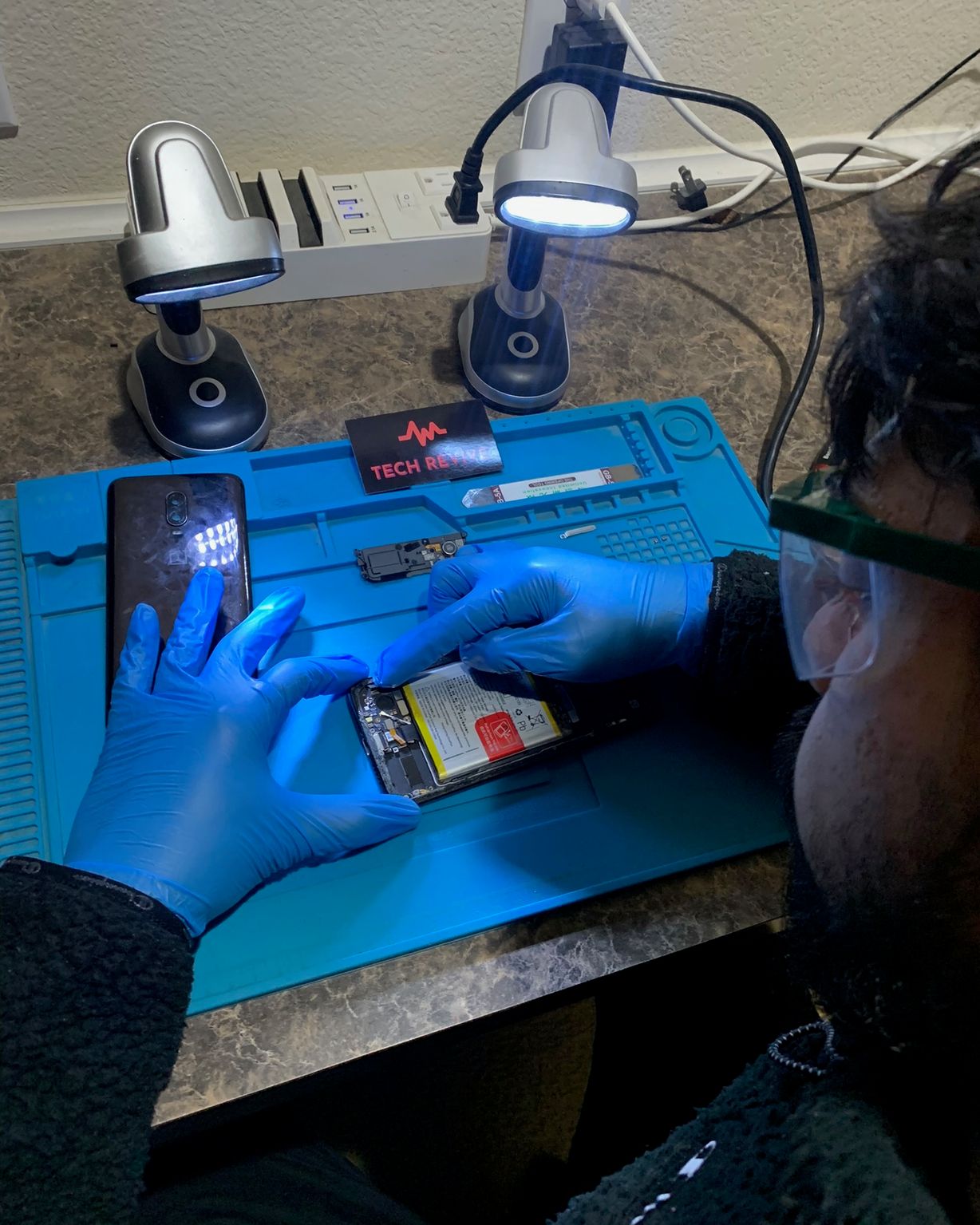 Tech Revive technician in safety glasses and blue gloves repairing a phone at a workbench